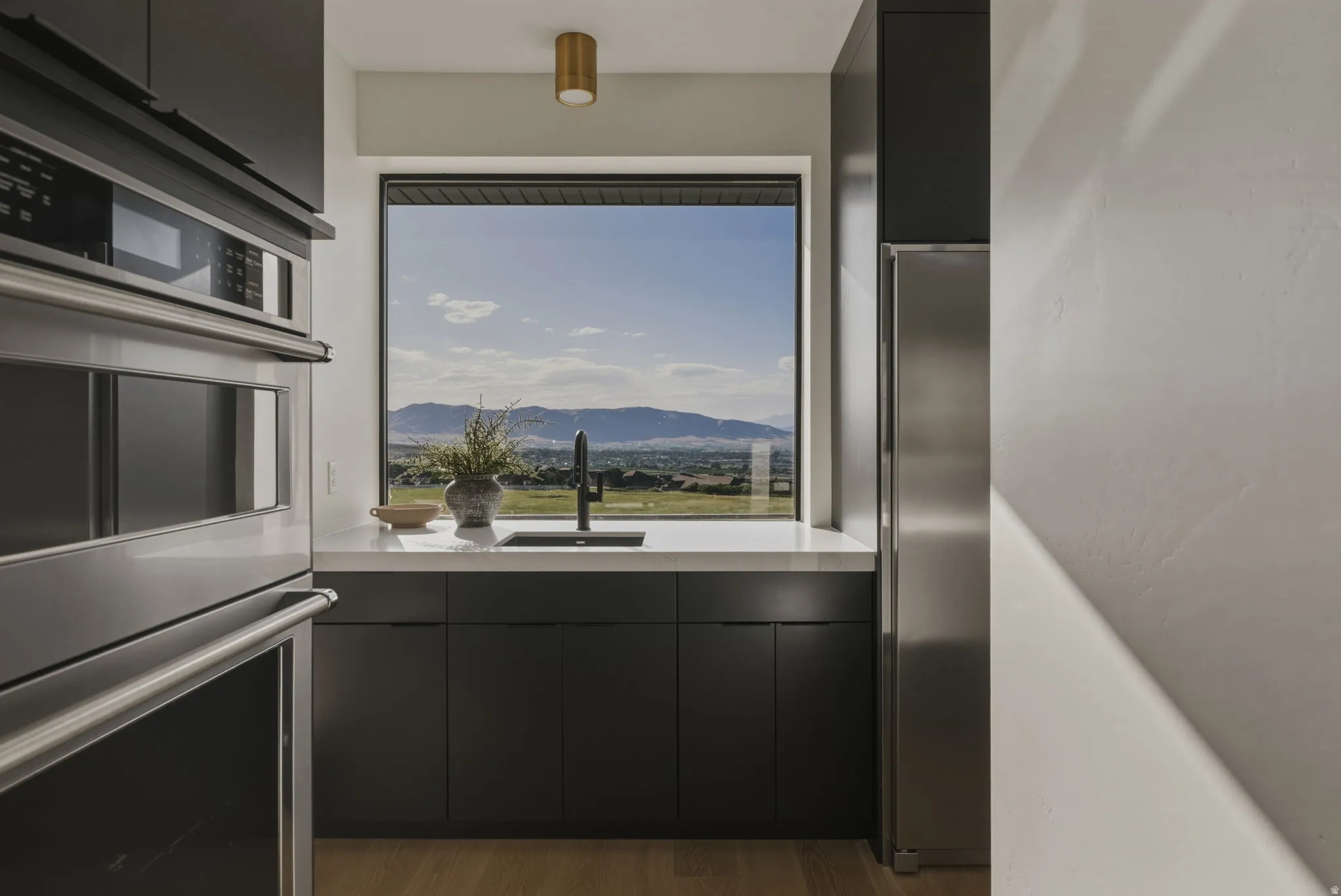 Modern kitchen featuring a large picture window with mountain scenery, sleek black cabinetry, white countertops, built-in stainless steel appliances, and wood-finish flooring