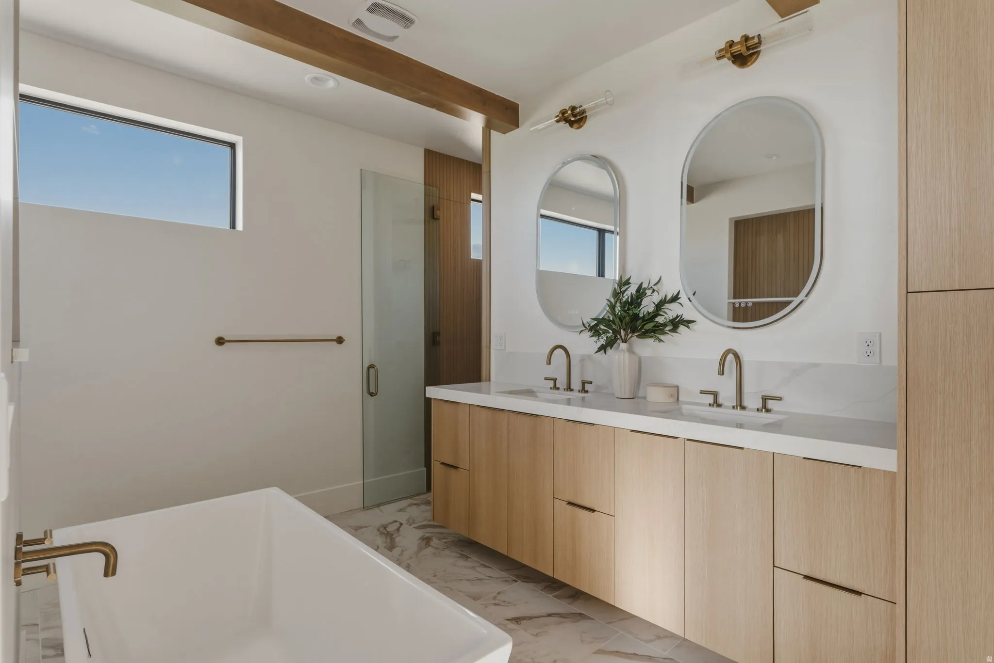 Contemporary bathroom featuring a light wood-finish double vanity with dual oval mirrors, a spacious freestanding soaking tub with a brass-finish faucet, and a glass-enclosed shower