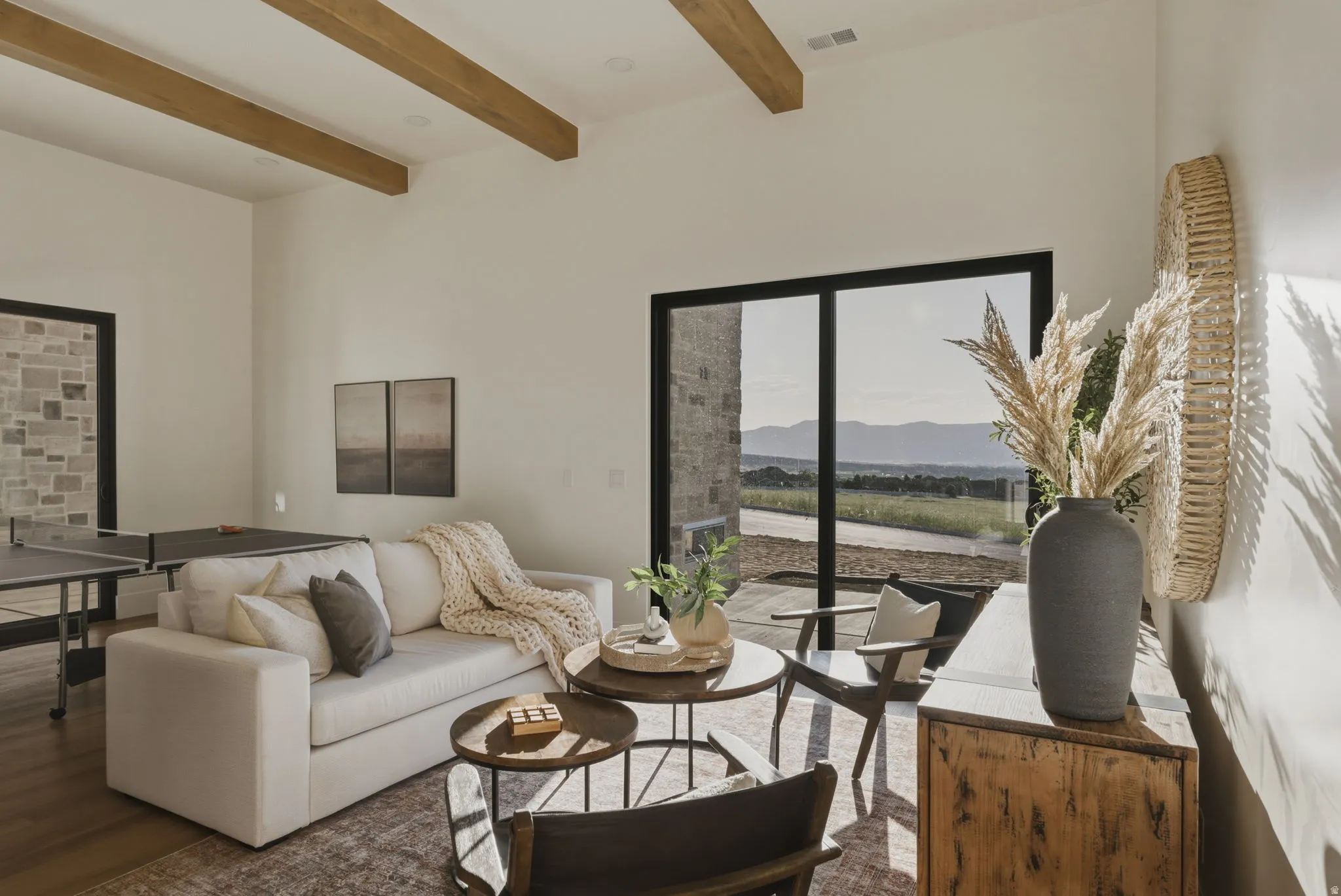Expansive living area featuring tall ceilings with exposed wood beams, wood-finish flooring, and a large sliding glass door opening to a mountain backdrop
