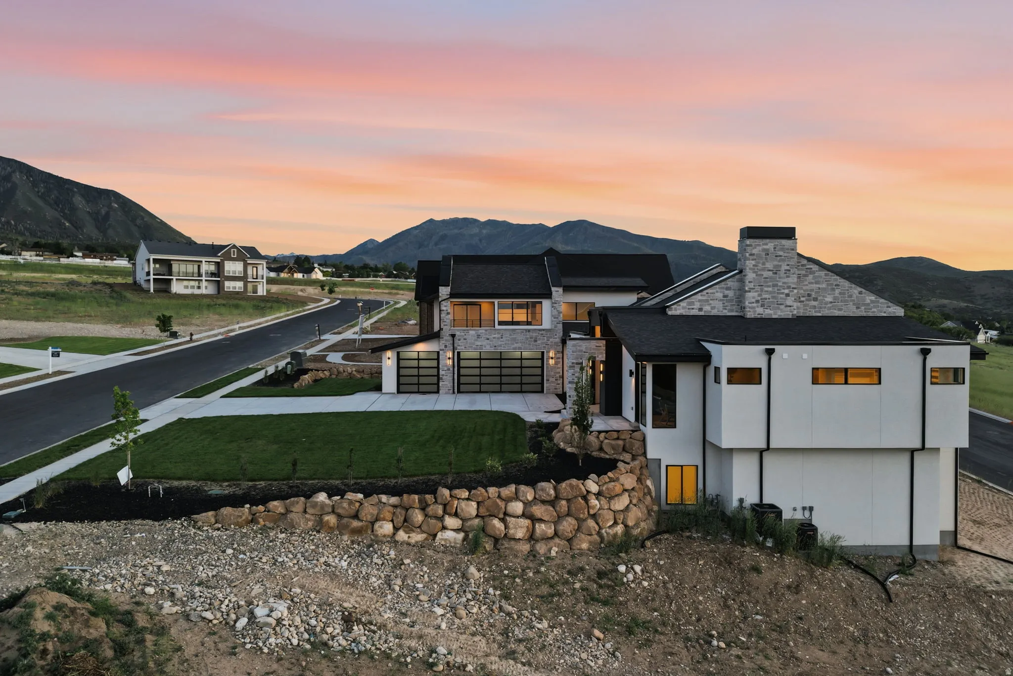 Modern architectural design featuring a stone and stucco exterior, black-framed garage doors, a stone retaining wall, and a black shingle roof