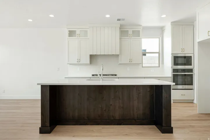 Modern kitchen featuring a dark wood-finish island with a light-toned countertop, white cabinetry with glass-front uppers, stainless steel built-in appliances, and light wood-finish flooring