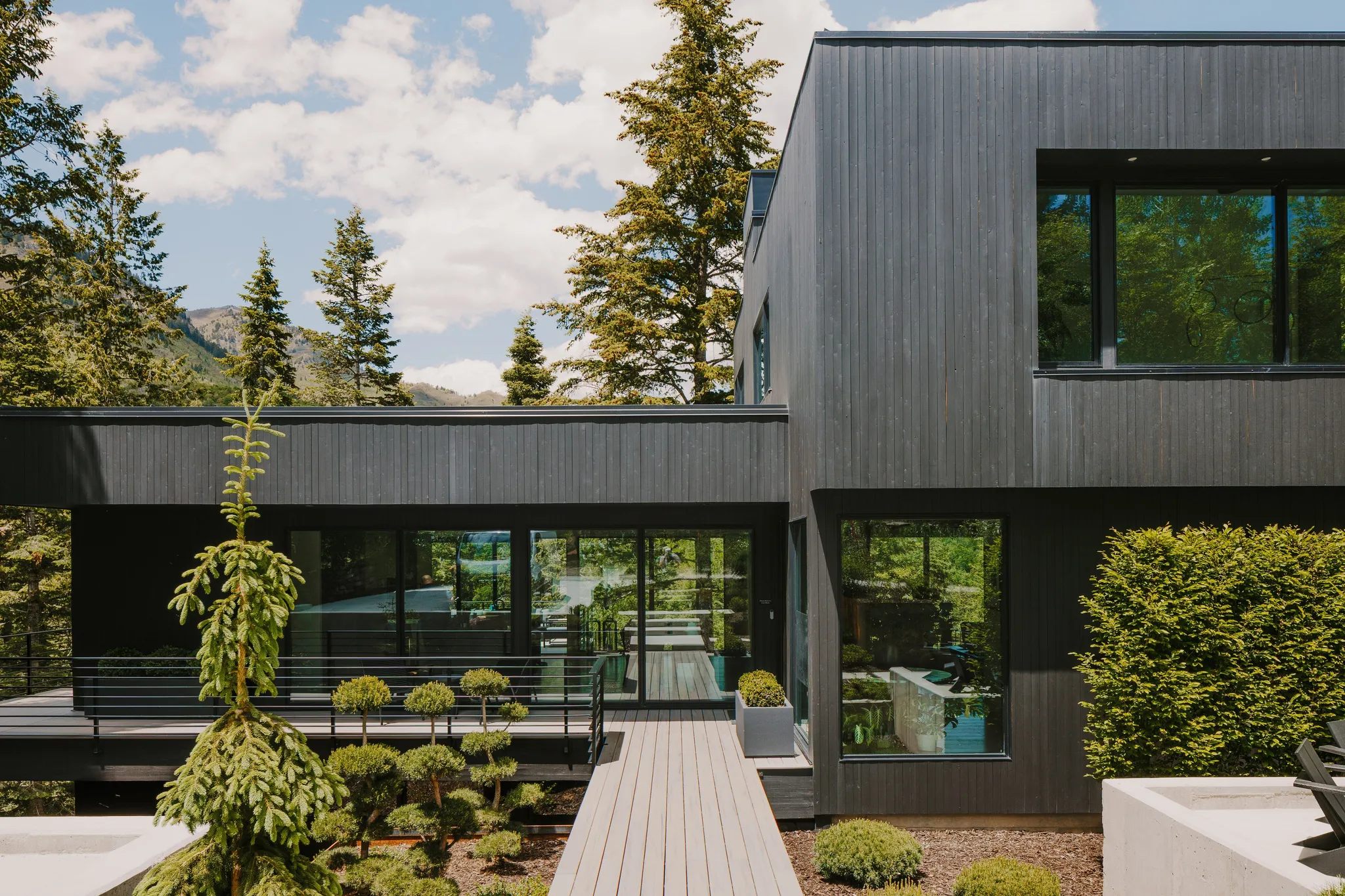 Modern exterior featuring black vertical siding, multiple large windows, a spacious deck with dark railings, a light-colored pathway, and diverse landscaping with mature trees and shrubbery