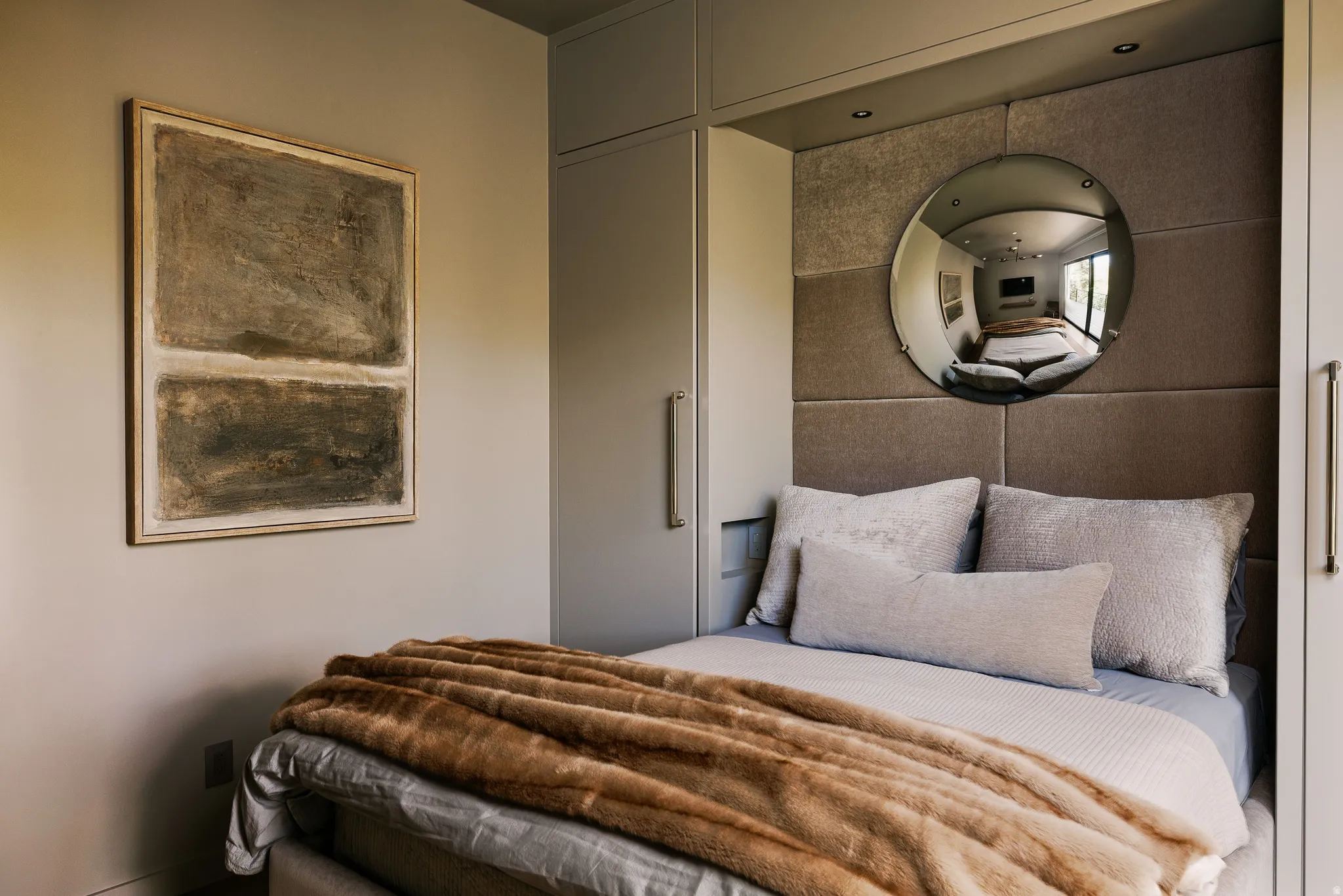 Built-in bed nook featuring a padded headboard with integrated lighting, a circular wall mirror, and flanking custom cabinetry with elongated hardware