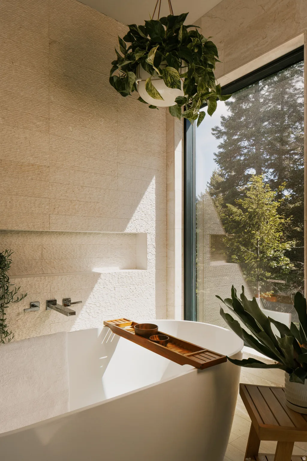 Soaking tub with wall-mounted faucet, textured stone tile walls, integrated shelving niche, and large picture window