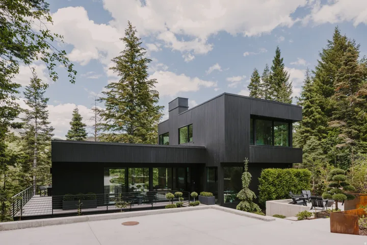 Contemporary architecture featuring a dark exterior finish, expansive windows, a concrete patio with built-in seating, a spacious outdoor deck, and mature evergreen landscaping