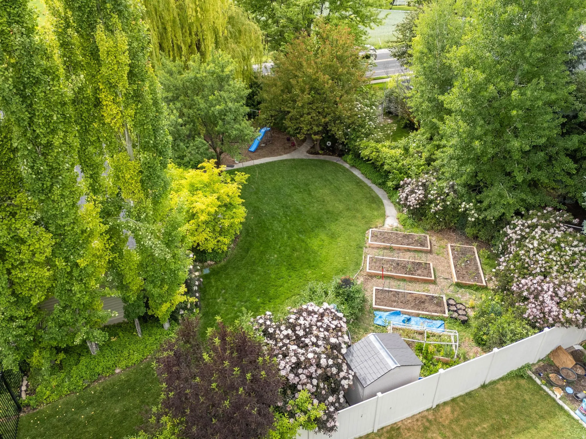 Expansive backyard featuring a manicured lawn, established garden beds, mature trees, a white privacy fence, and a storage shed