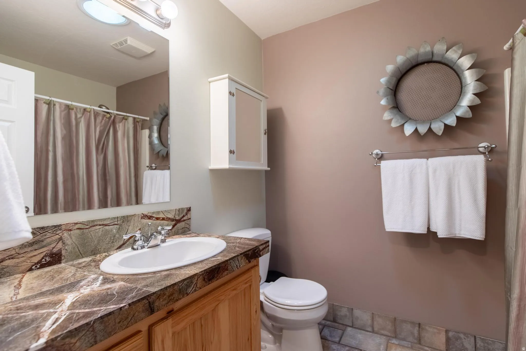 Bathroom featuring a light wood-finish vanity with a built-in sink, a patterned stone countertop, and a vanity mirror with overhead lighting