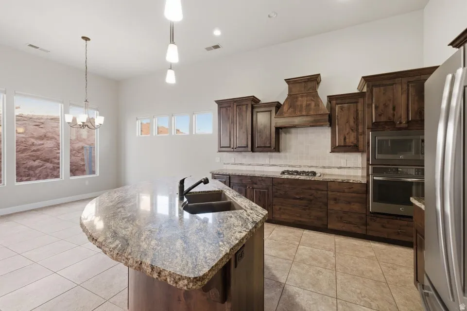 Spacious kitchen featuring an oversized island with a dual-basin sink and a natural stone countertop, wood-finish cabinetry, stainless steel appliances including a built-in oven and microwave, and a tile backsplash