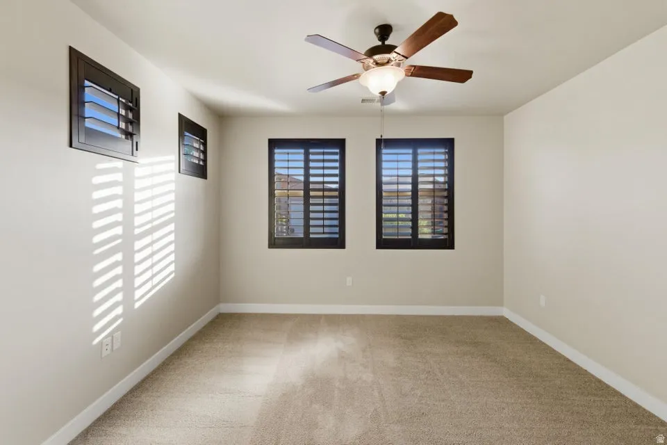 Neutral-toned carpeted room featuring three windows with dark-framed plantation shutters and a ceiling fan with light fixture