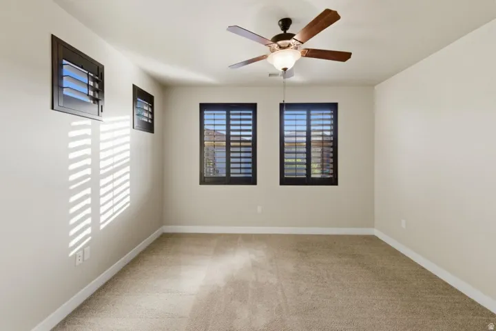 Neutral-toned carpeted room featuring three windows with dark-framed plantation shutters and a ceiling fan with light fixture