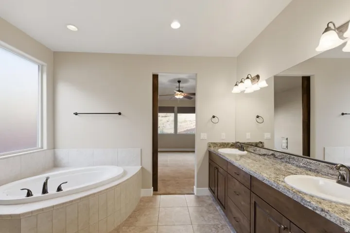 Bathroom featuring a large soaking tub with tile surround, dual vanity with undermount sinks and stone-finish countertop, dark wood-finish cabinetry, and tiled flooring