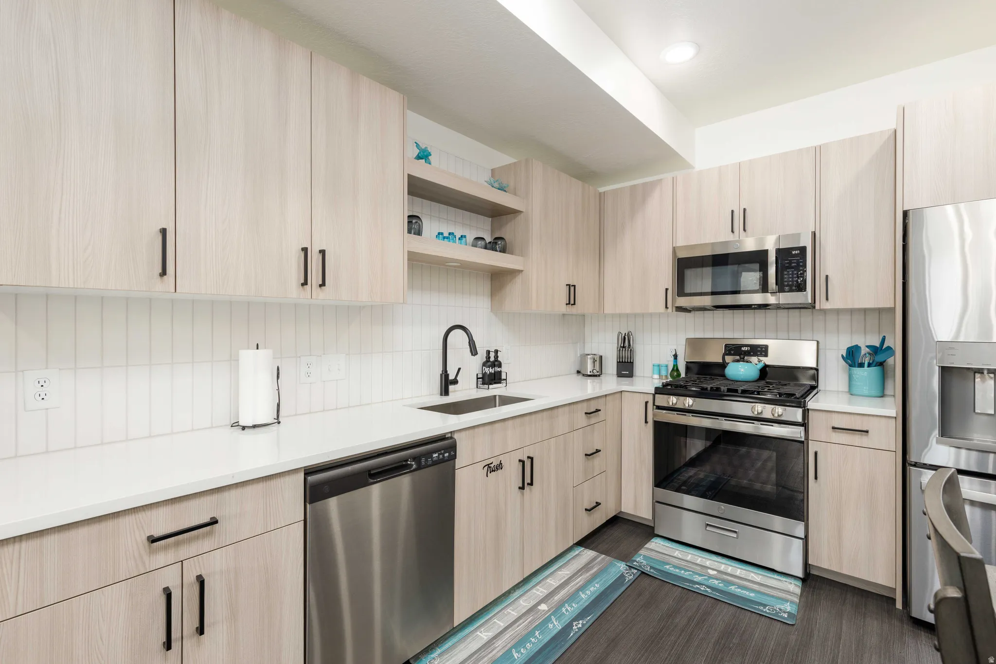 Modern kitchen featuring light wood-finish cabinetry, white countertops, and a full suite of stainless steel appliances including a refrigerator, range, microwave, and dishwasher