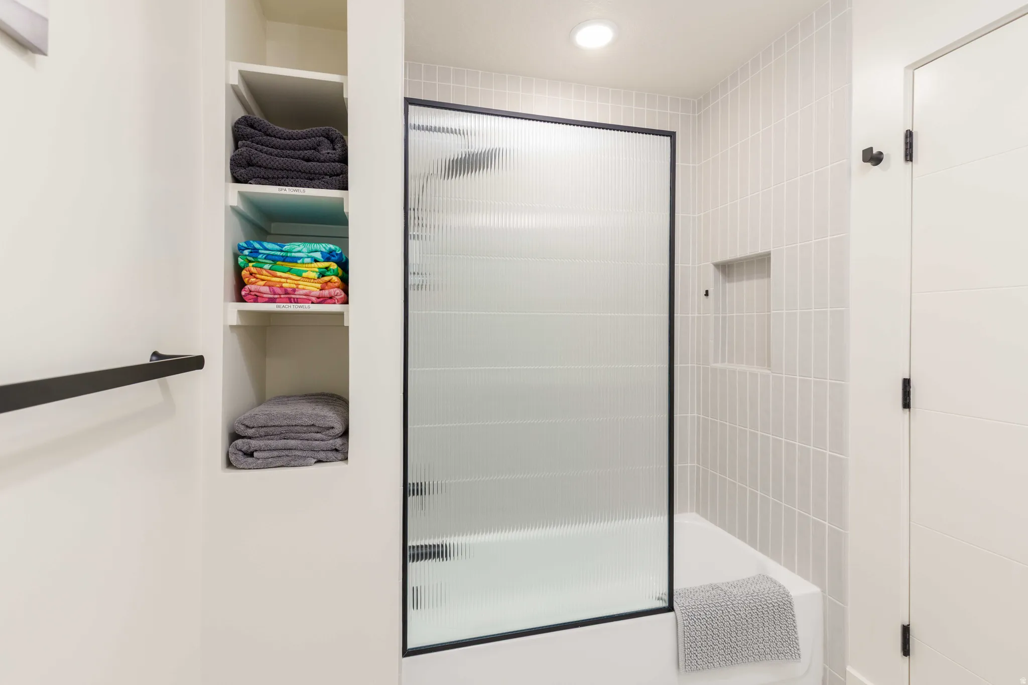Bathroom featuring a tub/shower combination with a fluted glass and black-framed shower door, light-toned vertical tile surround, and a recessed shower niche