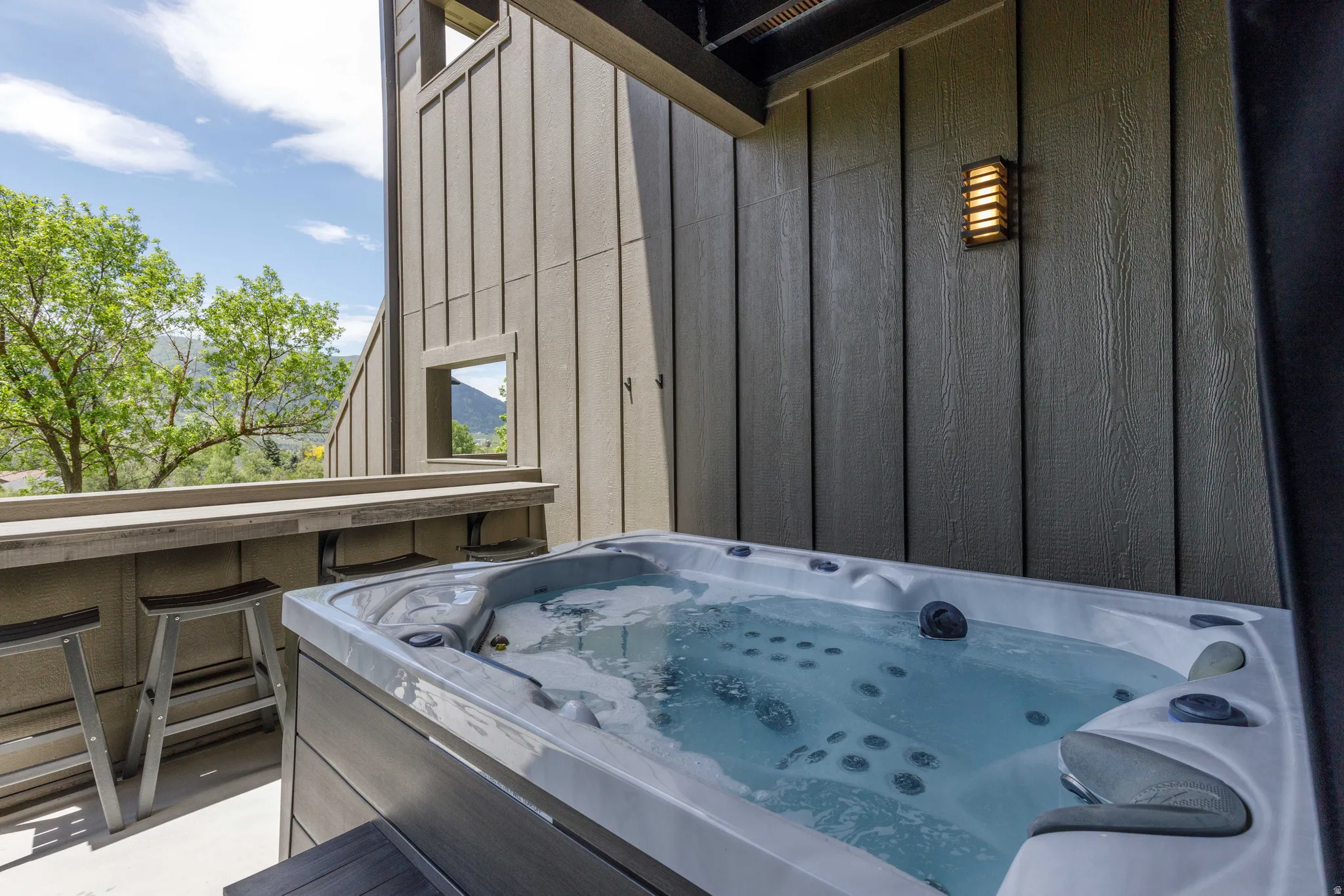 Outdoor hot tub situated on a concrete patio, featuring a built-in bar with seating, wood-paneled exterior walls, and a wall-mounted light fixture