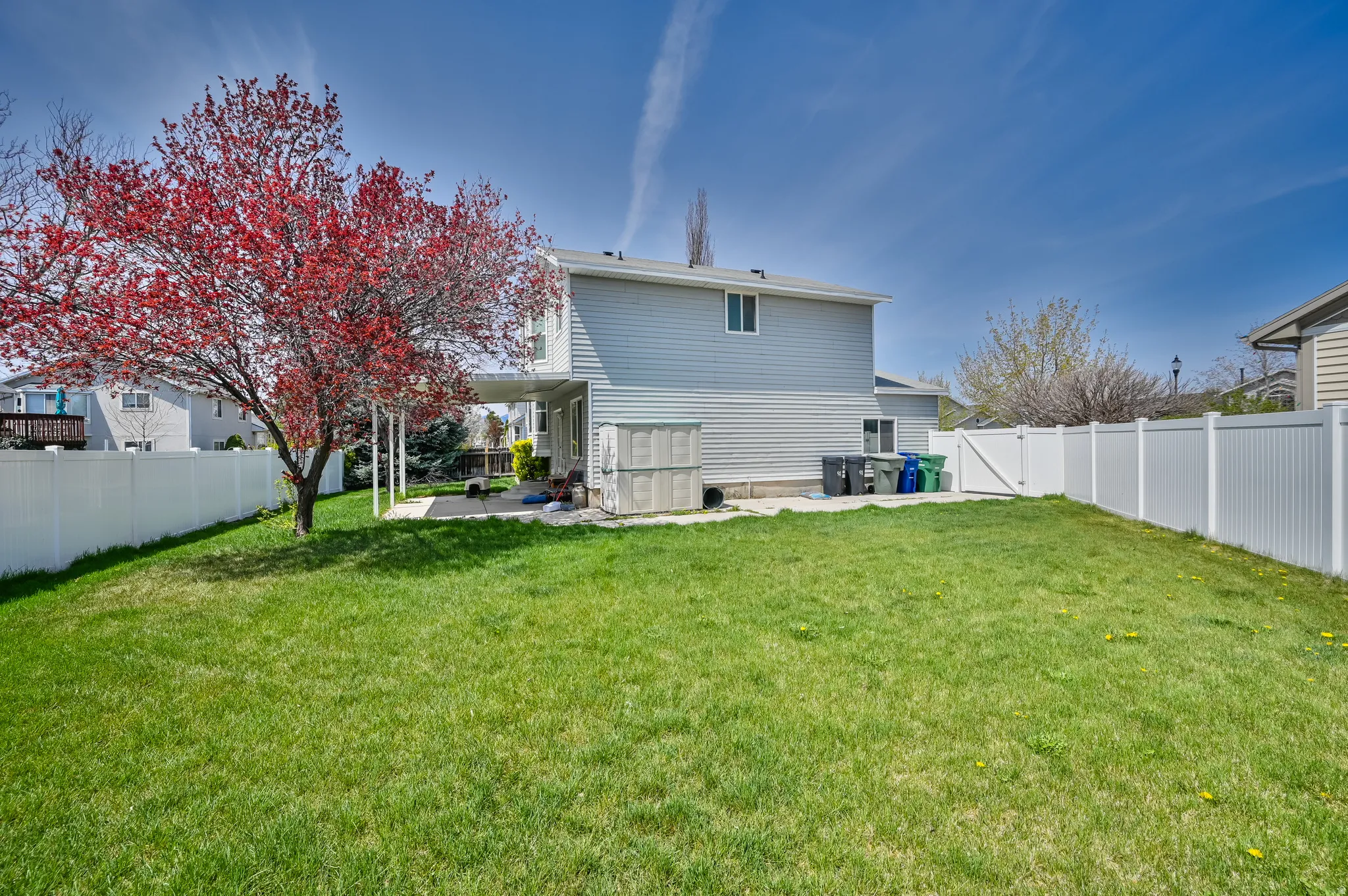 Expansive backyard featuring a manicured lawn, a mature red-leafed tree, a covered patio with a concrete surface, and white privacy fencing