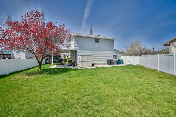 Expansive backyard featuring a manicured lawn, a mature red-leafed tree, a covered patio with a concrete surface, and white privacy fencing
