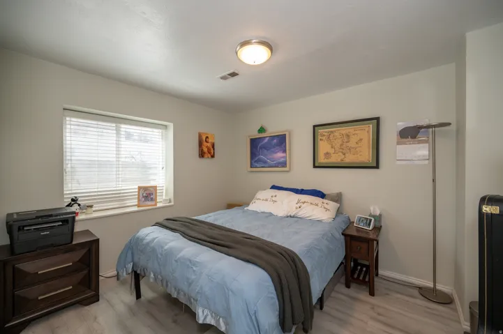 Bright bedroom featuring a large window with blinds, light-toned walls, overhead flush-mount lighting, and wood-finish flooring