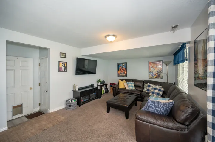 Carpeted living area featuring a mounted television above a media console, white painted walls, and a recessed window with blinds