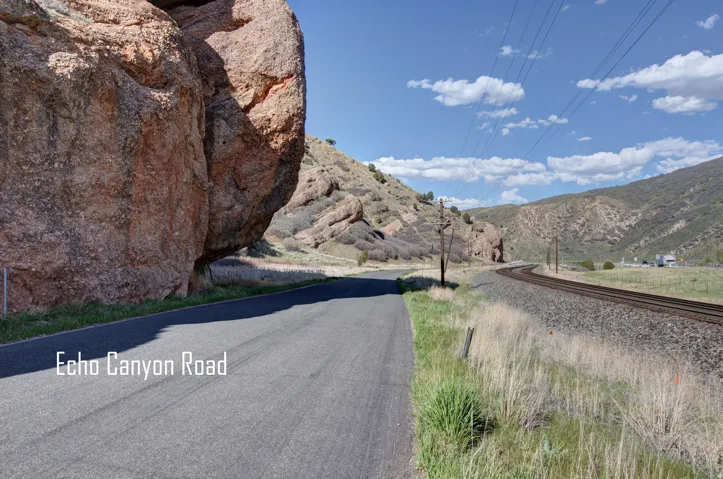 Paved road bordered by natural rock formations, featuring expansive mountain views, a railway line, and overhead power lines