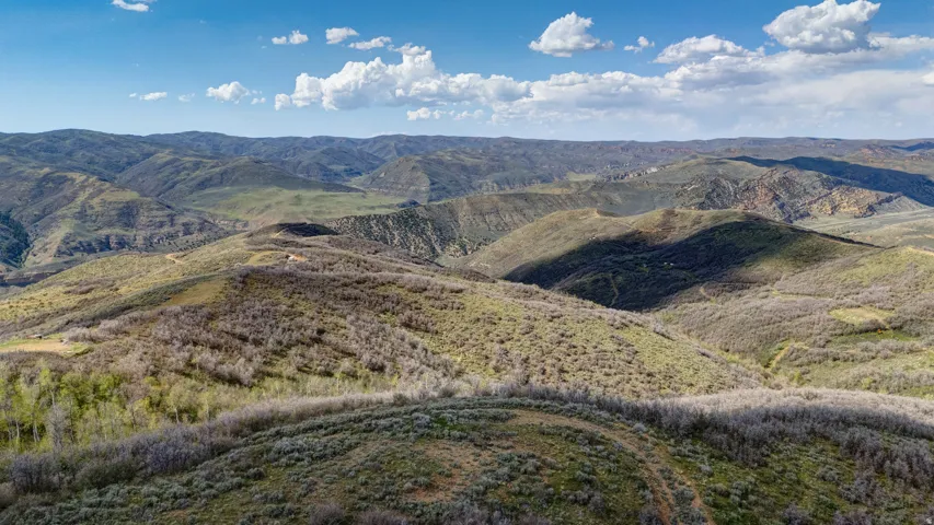 Expansive mountain landscape featuring rolling hills, varied green and brown vegetation, and a clear blue sky with scattered clouds