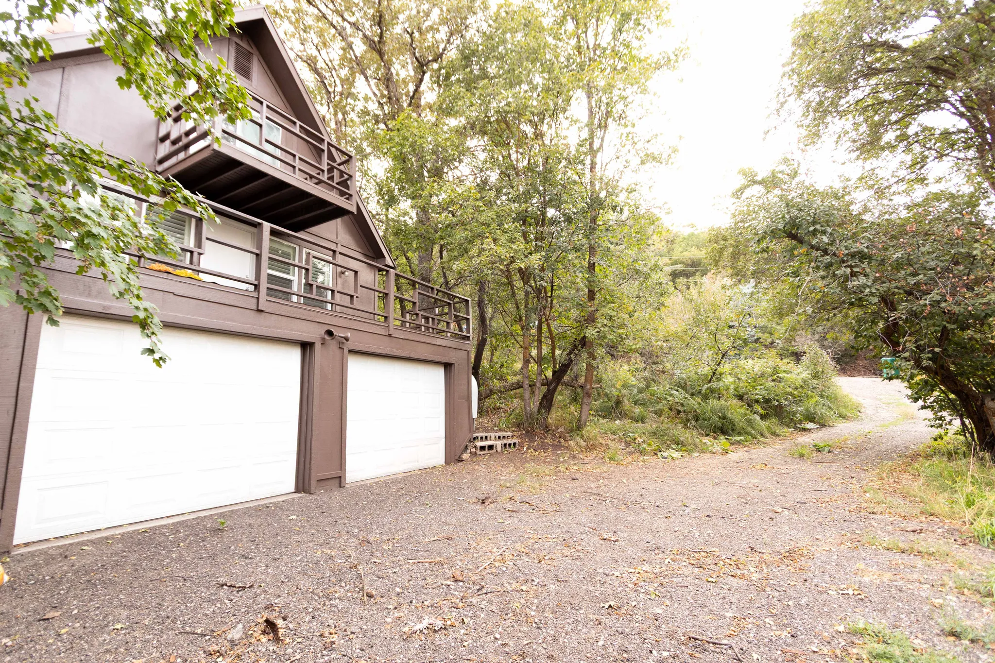 A-frame exterior with two balconies, dual garage doors, and a gravel driveway surrounded by mature trees