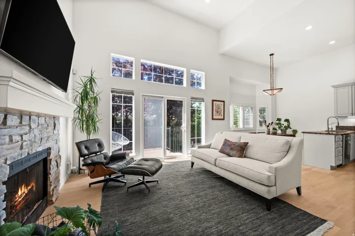 Vaulted ceiling living area featuring a stone-faced fireplace with a white mantel, maple hardwood floors (2024), and sliding glass doors to backyard deck & patio with hot tub.