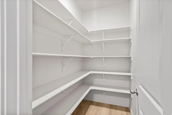 Walk-in pantry featuring extensive white shelving, wood-finish flooring, and a contemporary door with a brushed nickel knob