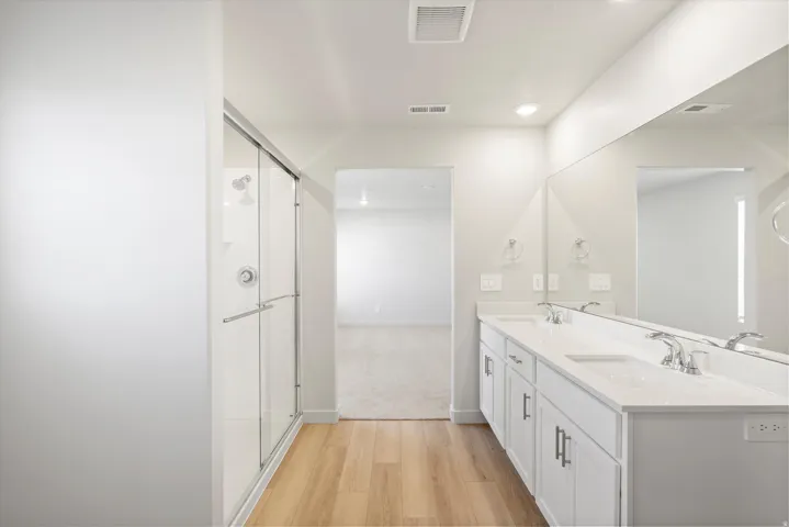 Spacious bathroom featuring a double vanity with white cabinetry and integrated sinks, a wide mirror, a glass-enclosed shower, and wood-finish flooring