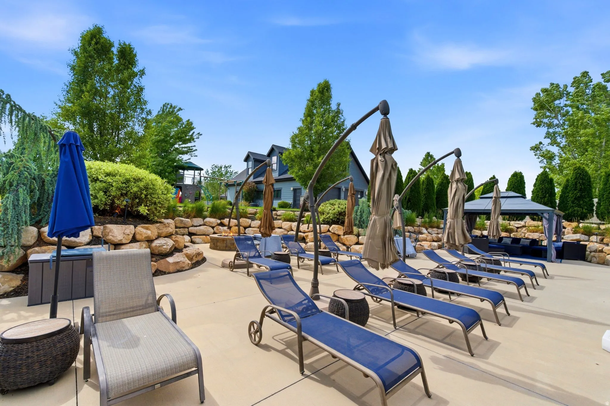 Expansive concrete patio with multiple lounge chairs, a fire pit, a covered gazebo seating area, and mature landscaping
