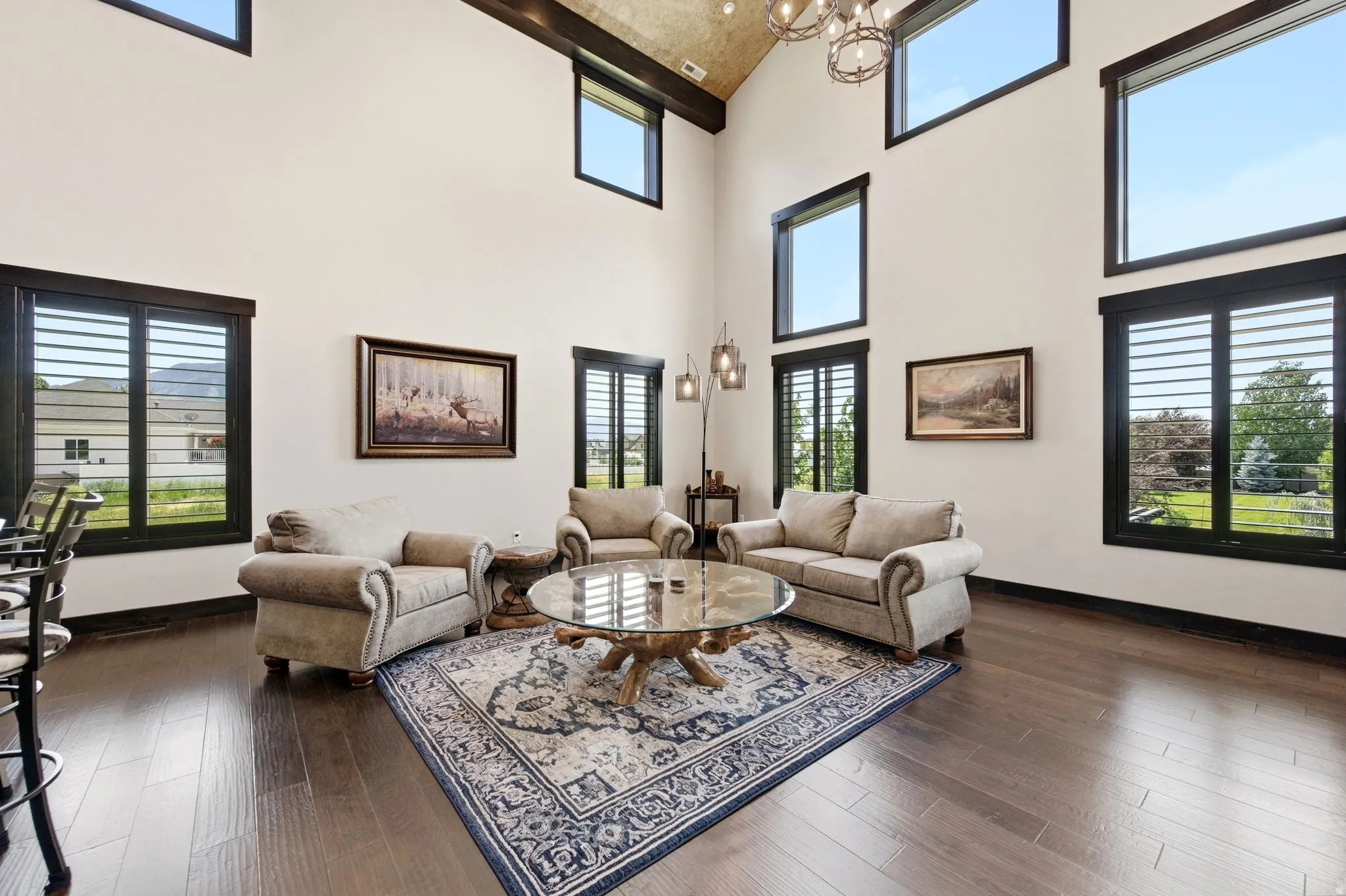 Spacious living area featuring a vaulted ceiling with exposed dark wood beams, wood-finish flooring, and multiple large windows with plantation shutters