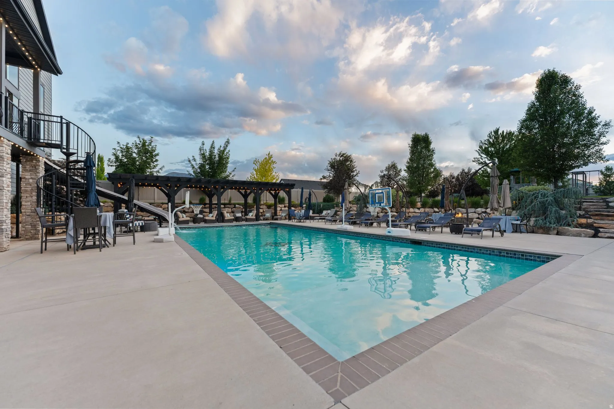 Expansive backyard featuring a rectangular swimming pool with brick coping, a black spiral staircase, and a dark wood pergola with string lights