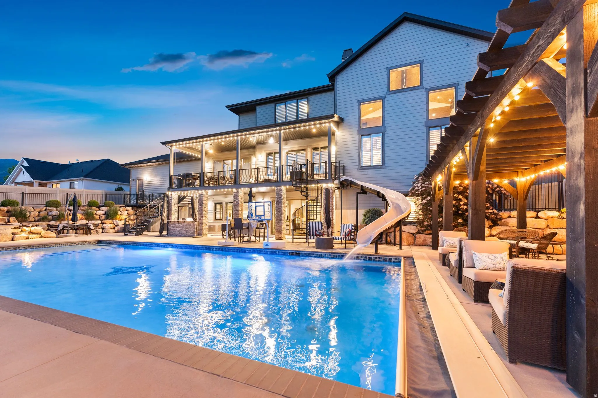 Expansive backyard featuring a large swimming pool with a slide, a two-story outdoor living area with a covered patio and balcony, and a pergola-covered seating area