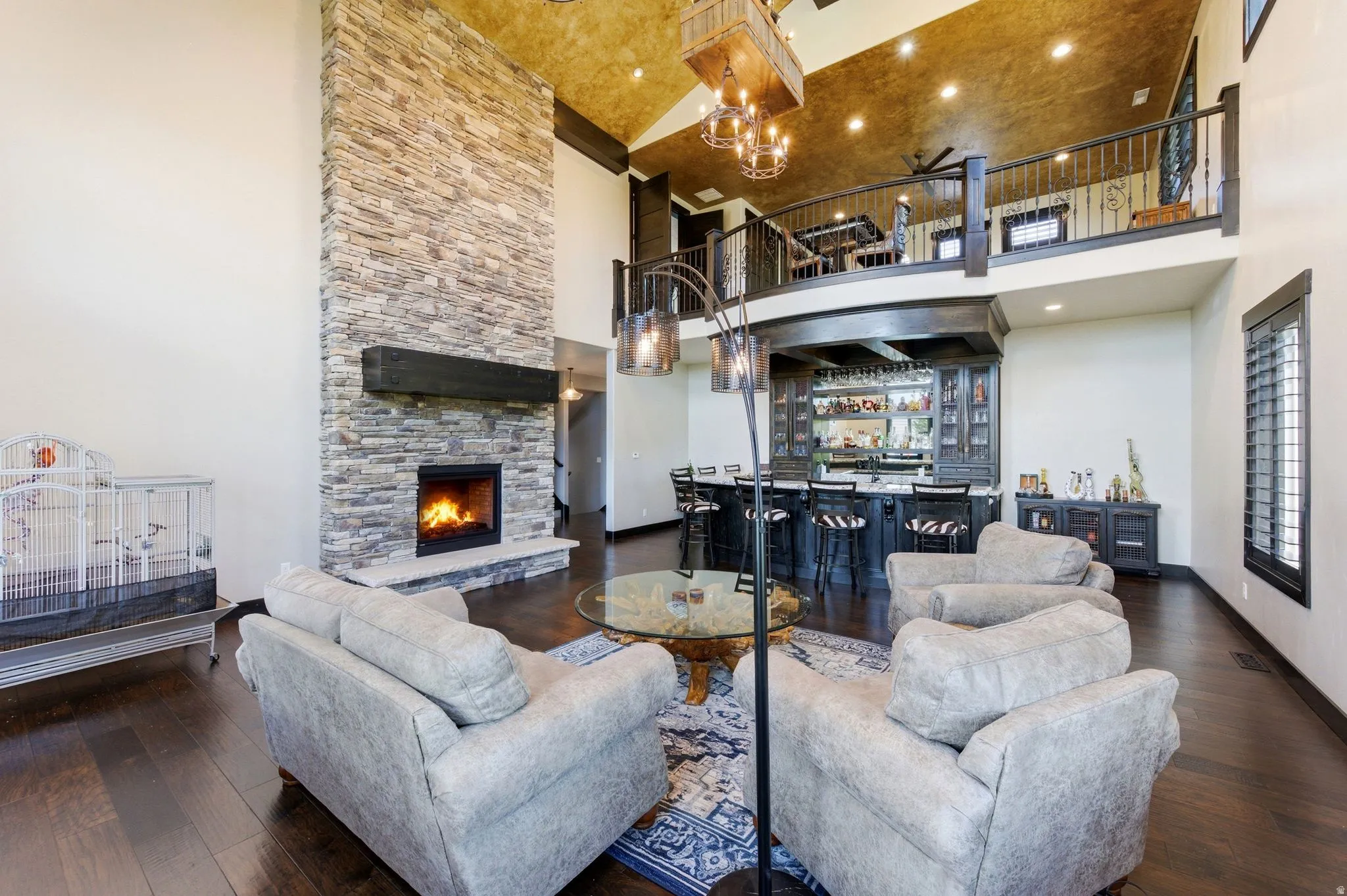 Two-story stone fireplace with hearth, dark wood-finish flooring, built-in wet bar with seating, upper-level balcony, and high vaulted ceiling with exposed beams
