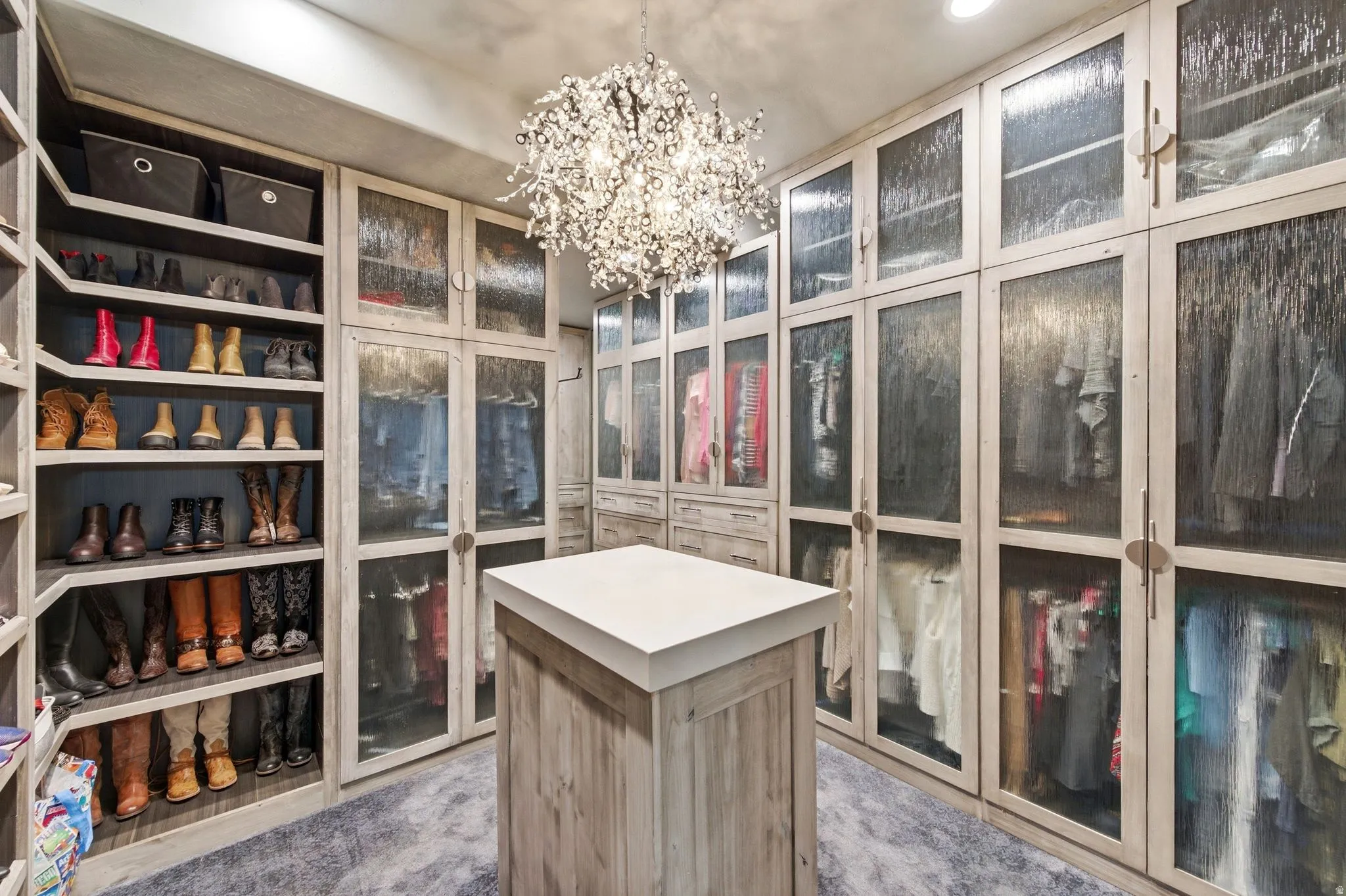 Custom walk-in closet featuring extensive built-in storage, textured glass cabinet doors, a central island with a solid surface countertop, wood-finish flooring, and a decorative chandelier