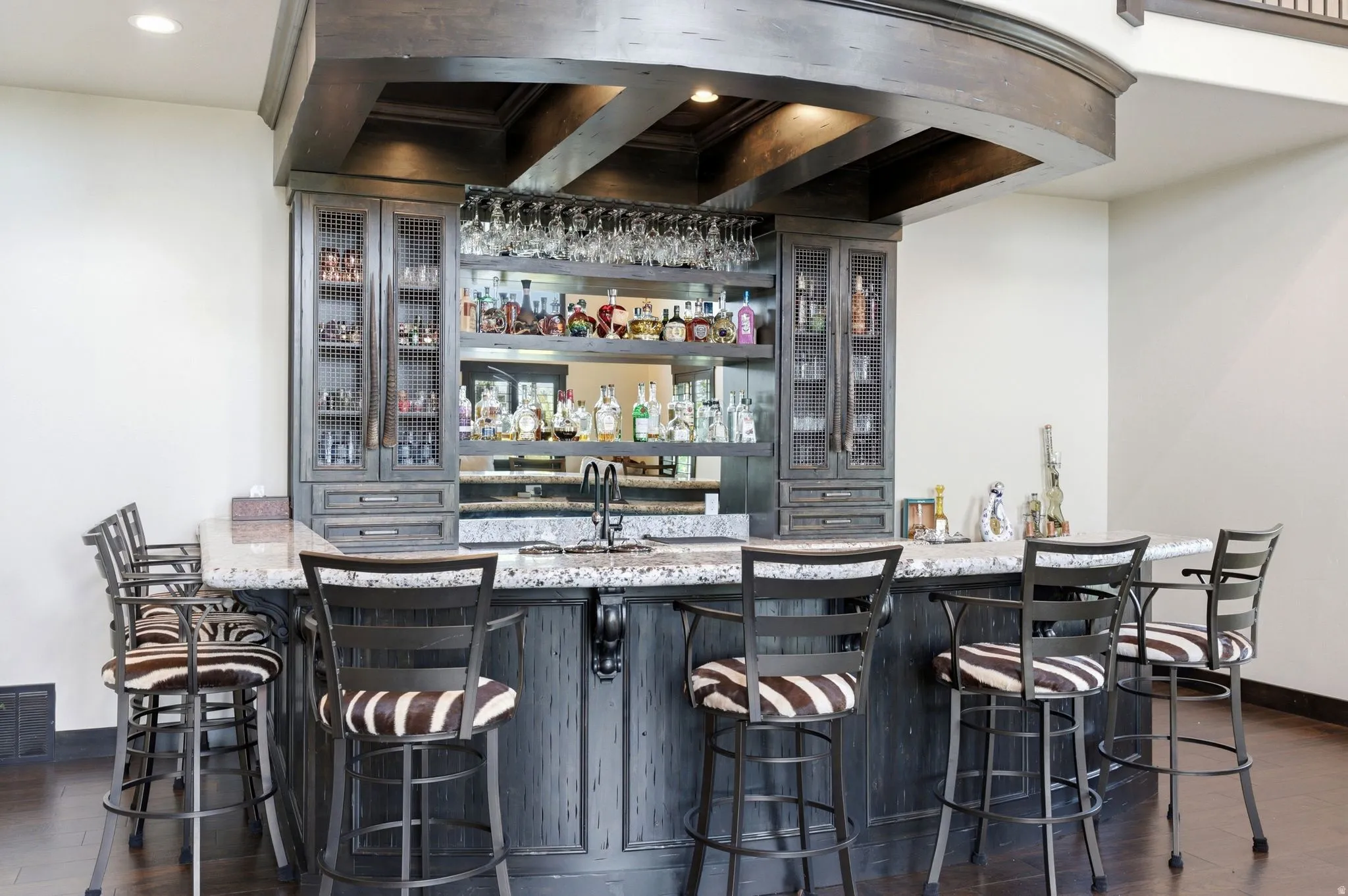 Built-in wet bar featuring a granite countertop, undermount sink with a gooseneck faucet, dark wood cabinetry with glass-front uppers, and integrated shelving for glassware