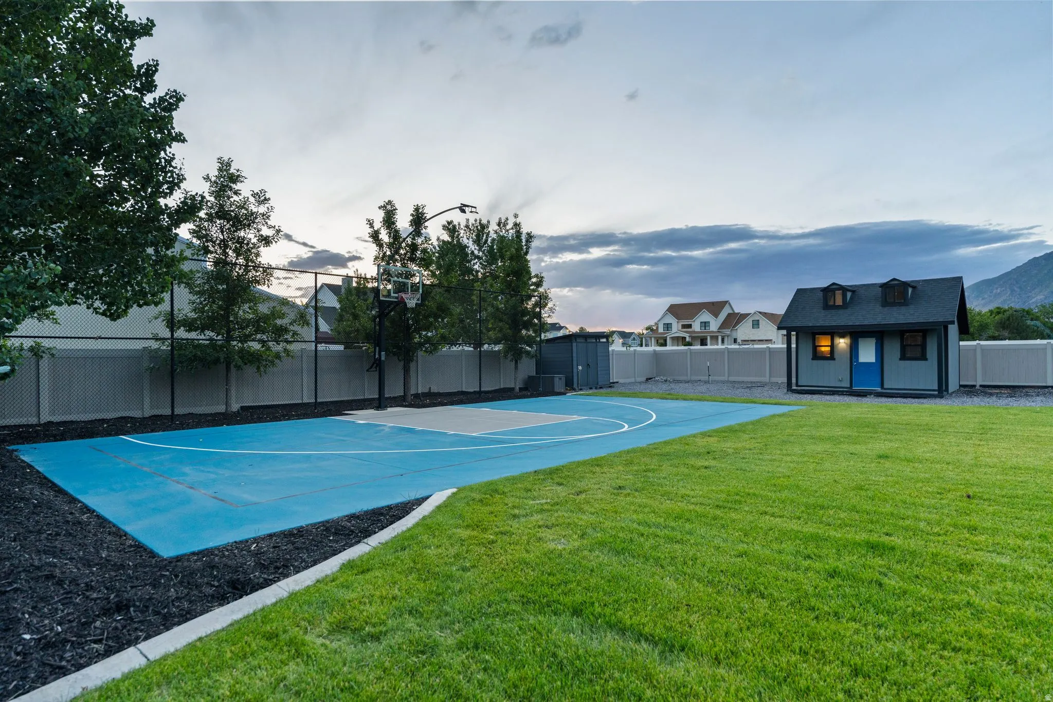 Expansive backyard featuring a blue-painted sports court with a basketball hoop, a manicured lawn, and a charming grey shed with a blue door and dormer windows