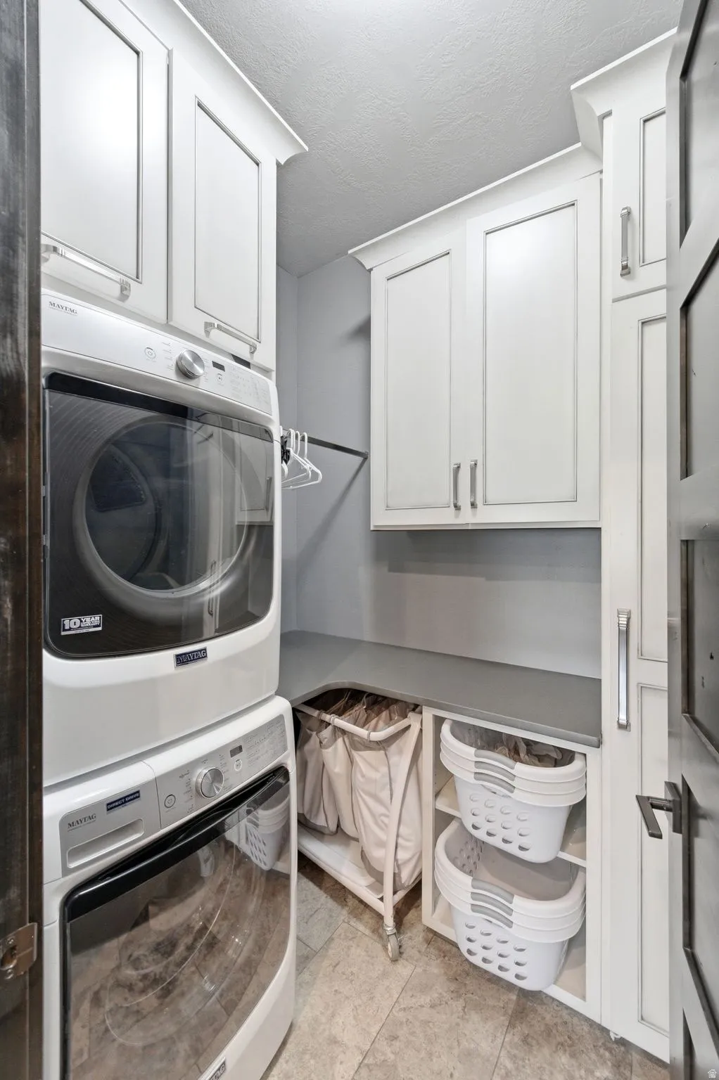 Dedicated laundry area featuring stacked appliances, ample white cabinetry with crown molding, a built-in hanging rod, a functional countertop, and tile flooring