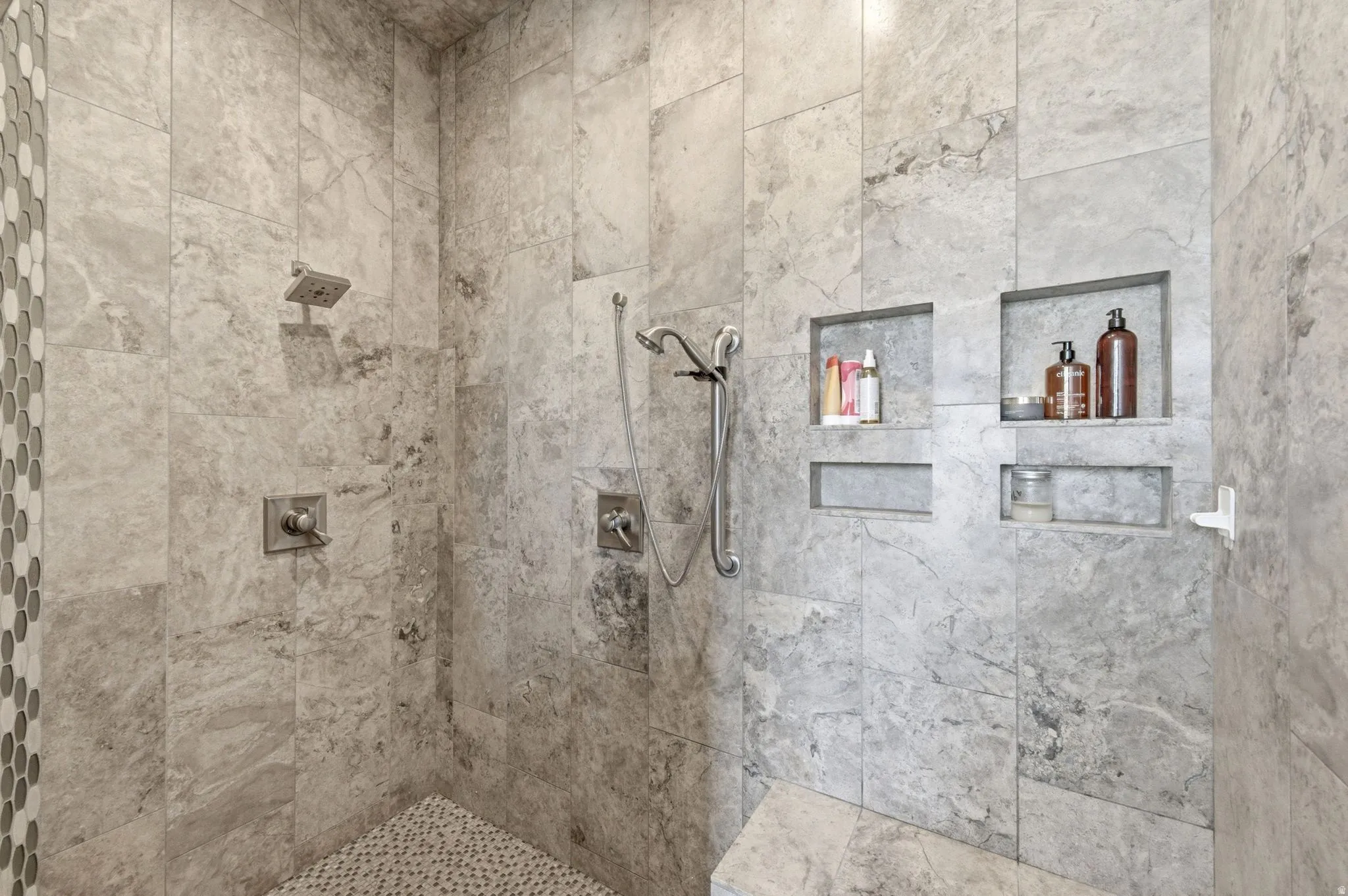 Spacious shower featuring floor-to-ceiling stone-finish tile, multiple recessed storage niches, a built-in shower bench, and dual shower heads