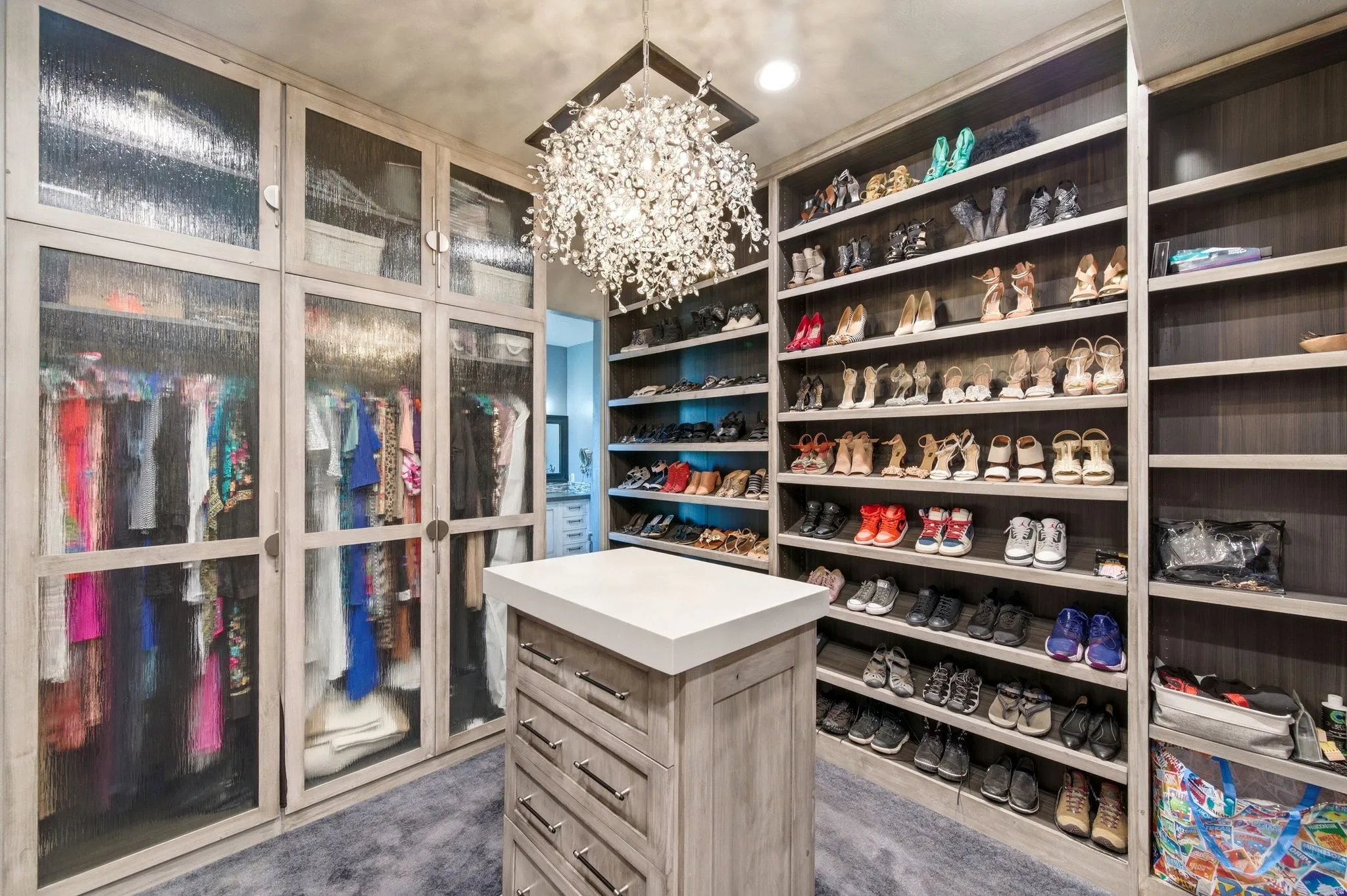 Custom closet featuring extensive open shelving, integrated cabinetry with textured glass doors, a central island with drawer storage and a light-toned countertop, and an overhead crystal chandelier