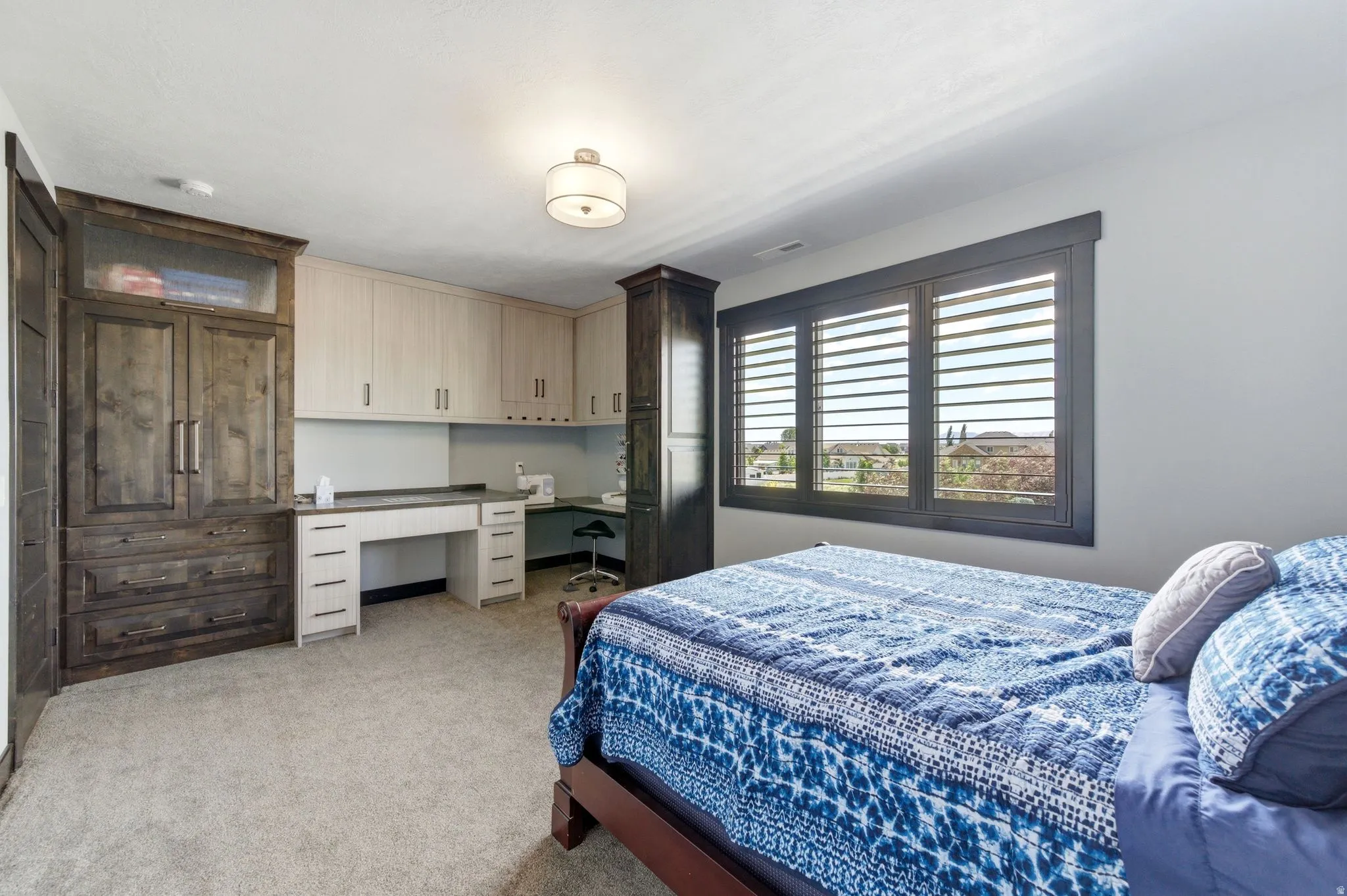 Room featuring textured carpet, a large window with dark-framed plantation shutters, a built-in desk area with light-toned cabinetry, and additional dark-toned built-in cabinetry with drawers and double doors