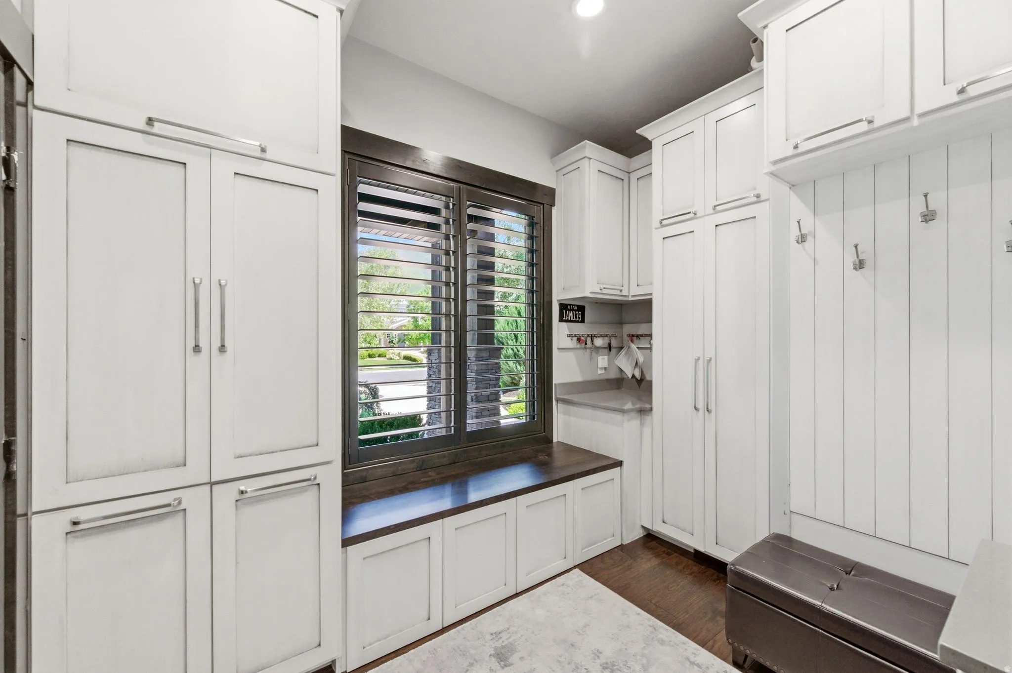 Custom white cabinetry throughout, dark wood-finish window trim with plantation shutters, built-in window bench, gray countertop, and wood-finish flooring