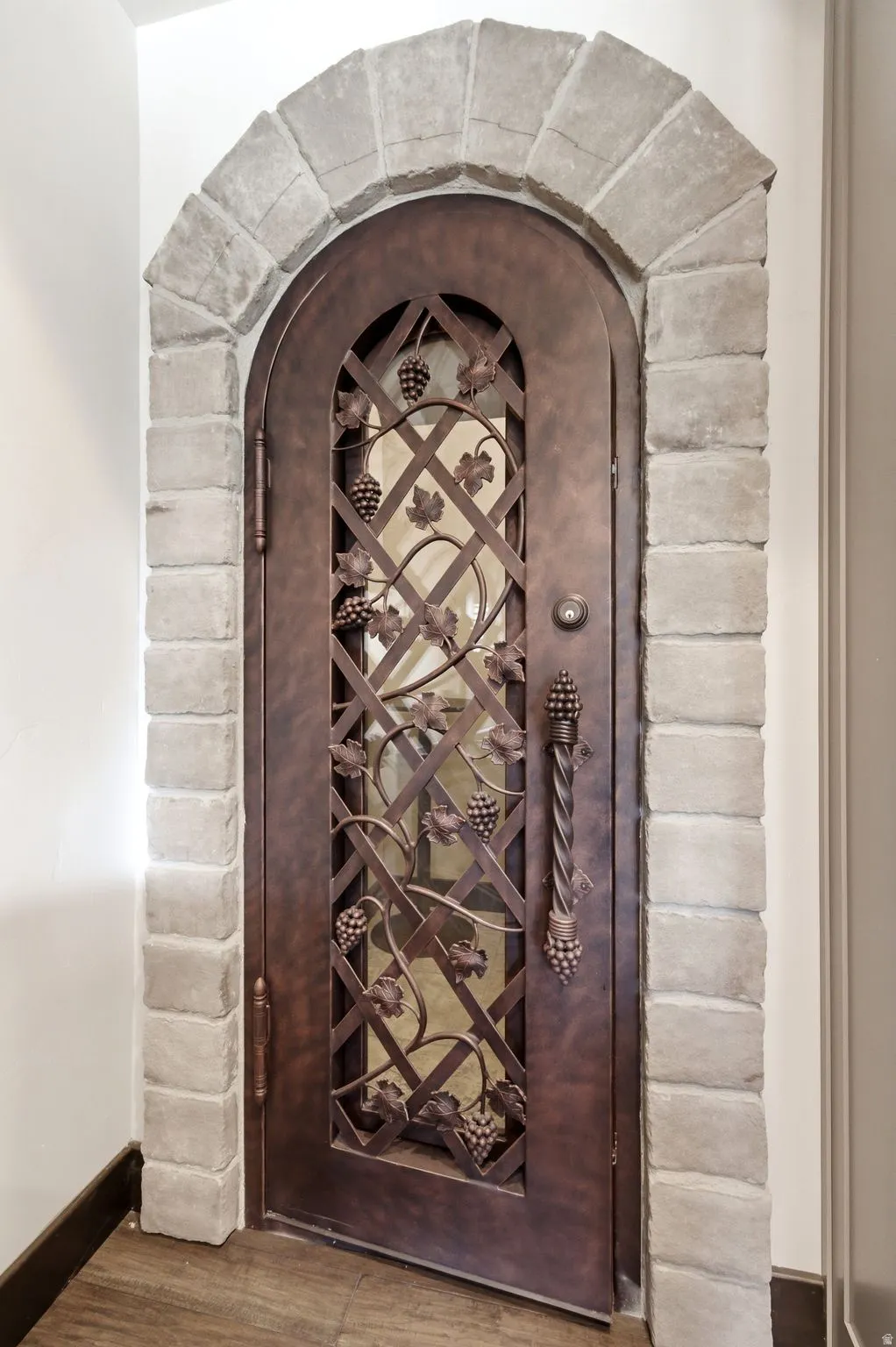 Arched stone doorway featuring a custom bronze-finish iron door with intricate grape and vine detailing, an ornate twisted handle, and obscured glass panels