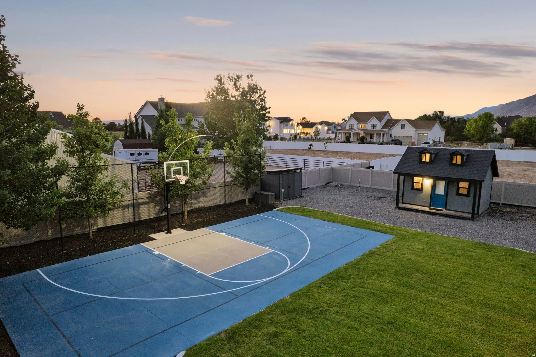 Expansive backyard featuring a blue-surfaced sports court with a basketball hoop, a dedicated green lawn area, a charming shed with dormer windows, a gravel-covered utility area, and a privacy fence