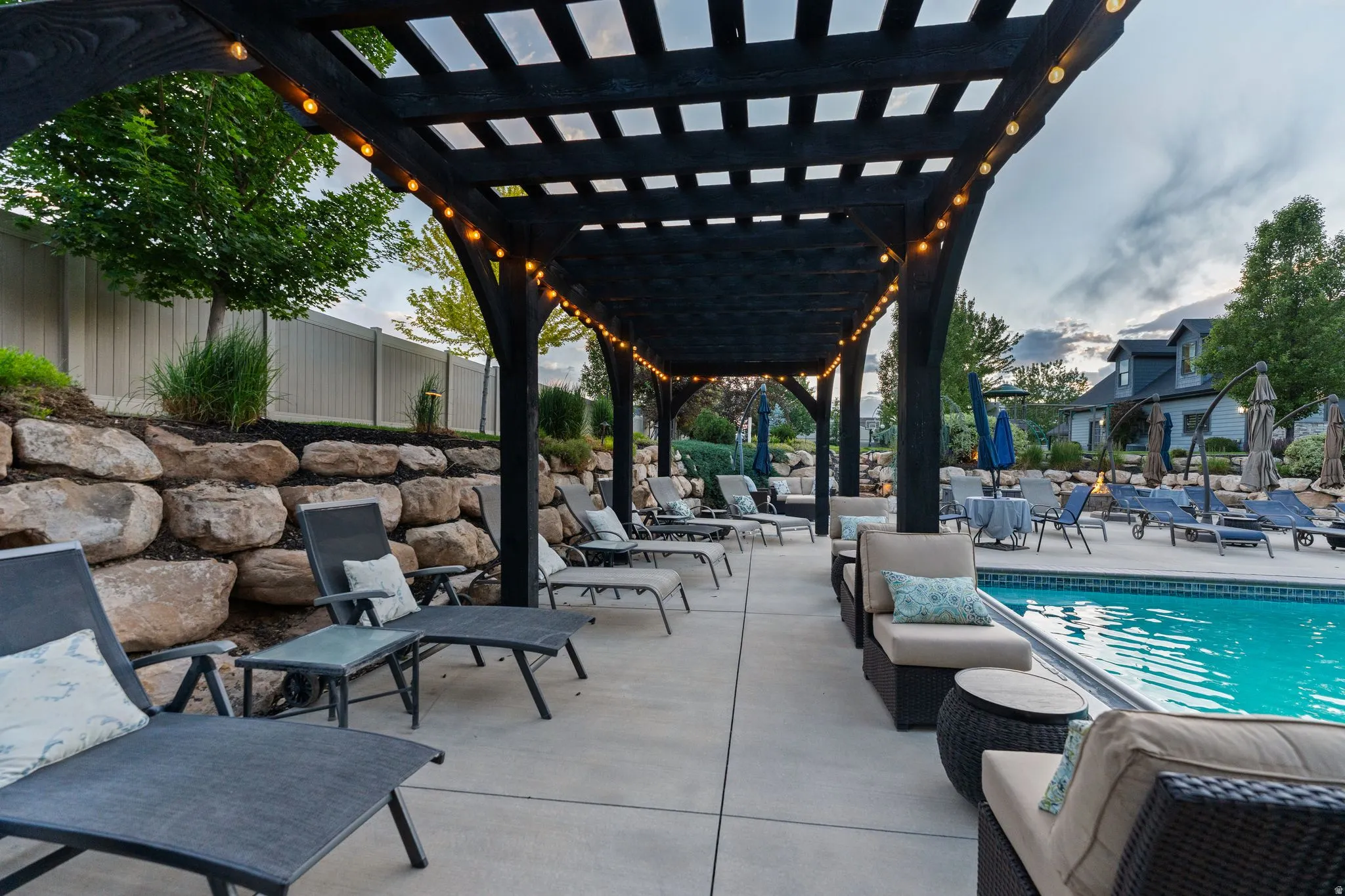 Expansive poolside patio featuring a pergola with integrated string lighting, concrete decking, and a rock retaining wall