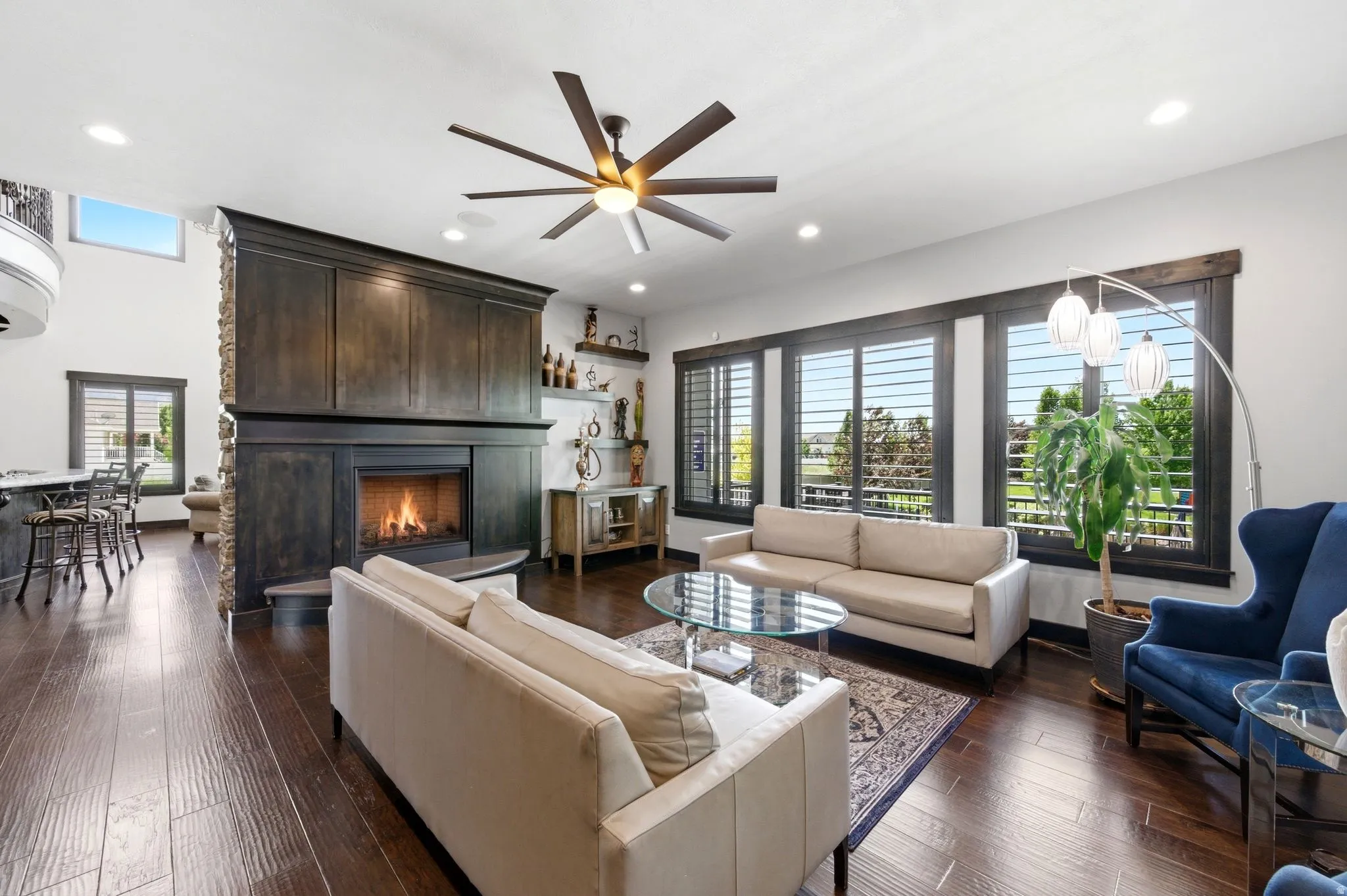 Spacious living area featuring rich wood-finish flooring, a stone-clad fireplace with dark wood mantel and cabinetry, and multiple large windows with plantation shutters