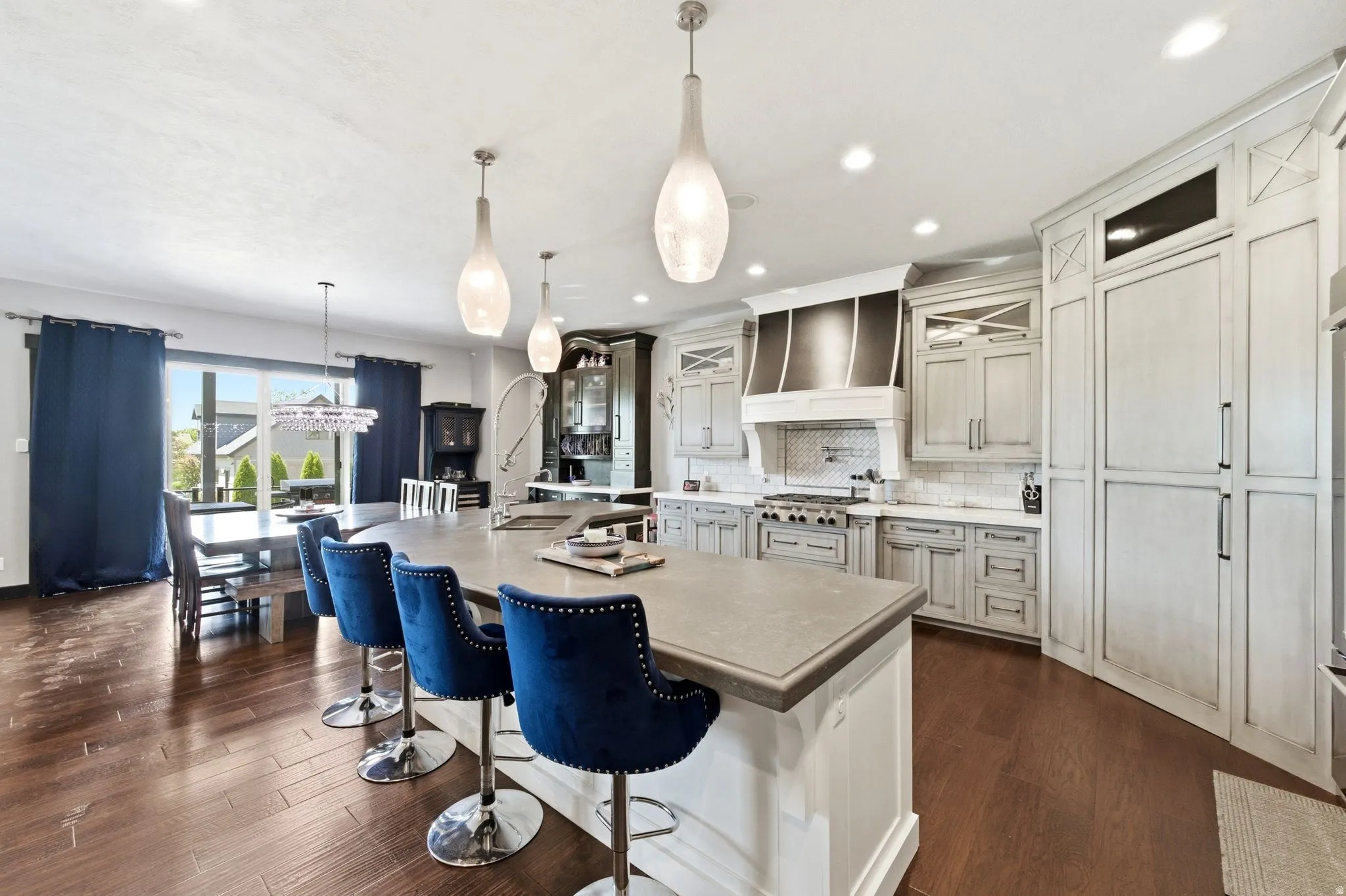 Gourmet kitchen featuring an oversized island with counter seating, light-toned custom cabinetry, wood-finish flooring, a tile backsplash, and stainless steel appliances including a range and range hood