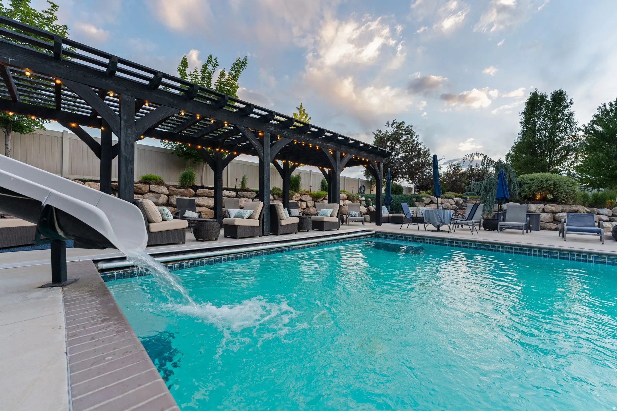 Outdoor swimming pool with a water slide, surrounded by a dark wood pergola with string lighting