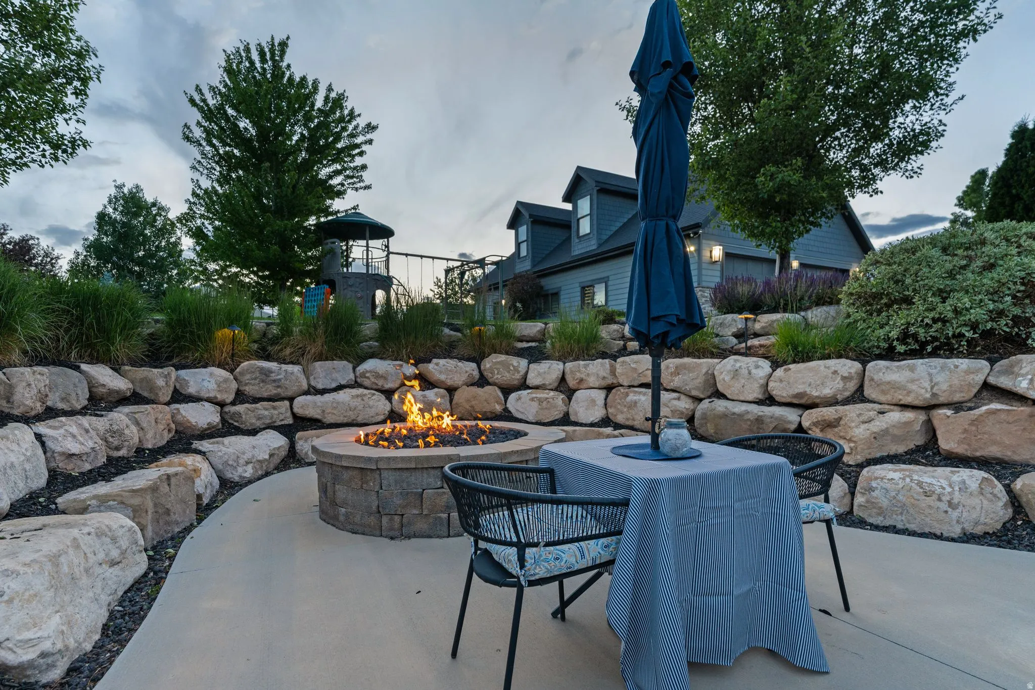 Outdoor patio featuring a stone fire pit, concrete surfacing, and a tiered rock retaining wall with landscaping