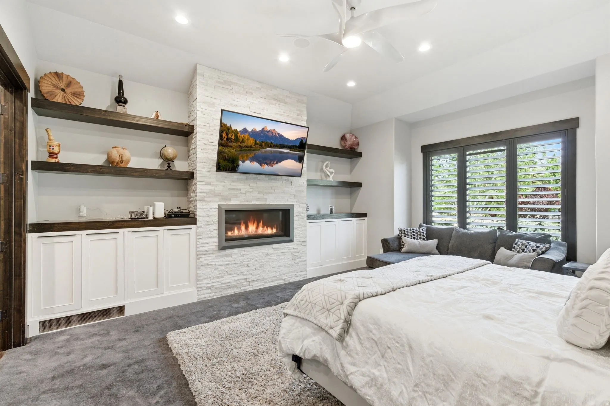 Carpeted room featuring a white stacked stone fireplace with an inset television, built-in cabinetry with wood-finish countertops and floating shelves, and a window with dark wood-finish shutters