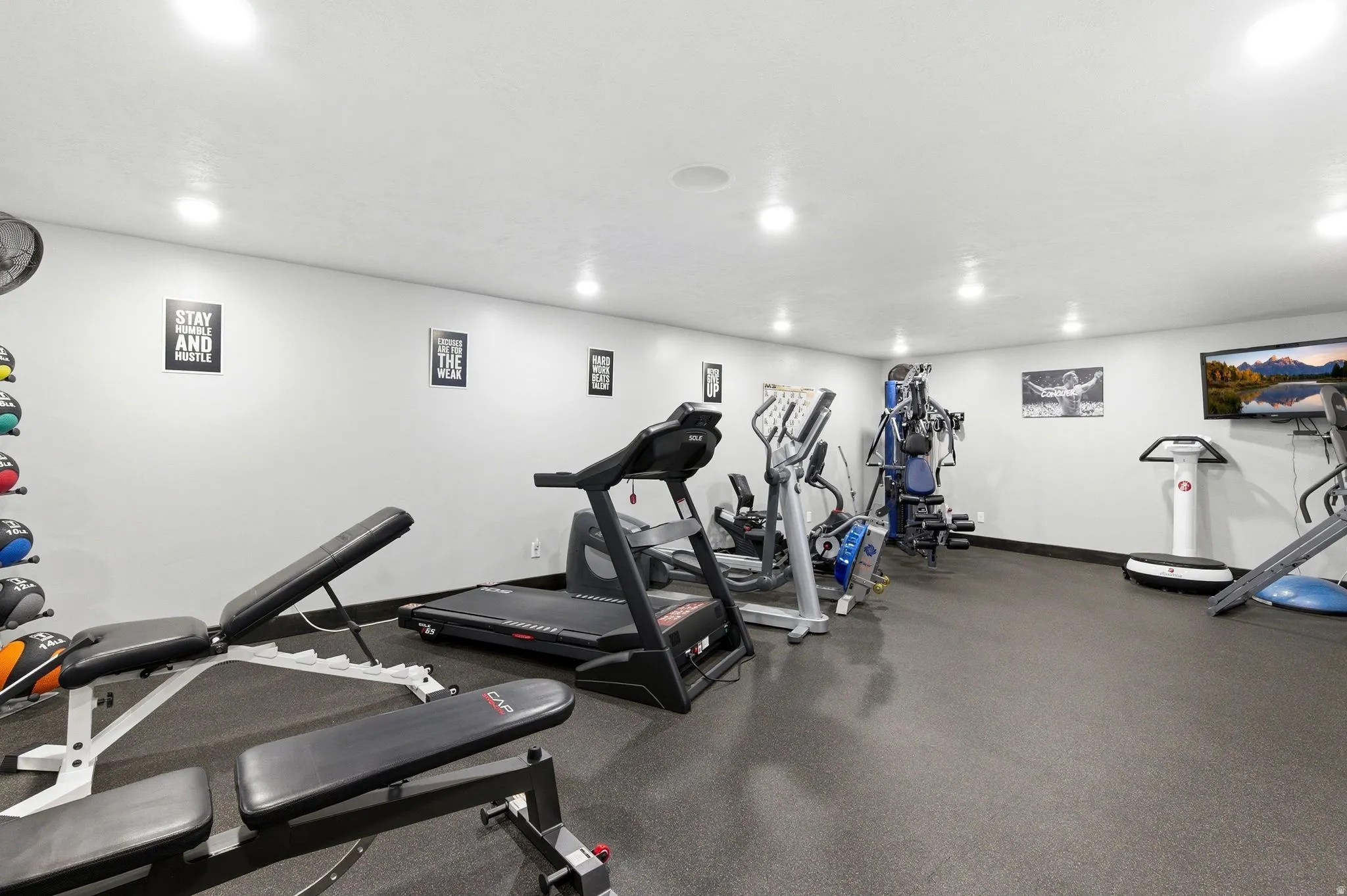 Dedicated fitness space featuring resilient black flooring, recessed ceiling lighting, a wall-mounted display, and bright white walls